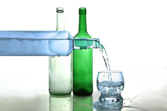 Dvě stojící lahve, ze třetí je nalévána do skleničky čirá tekutina